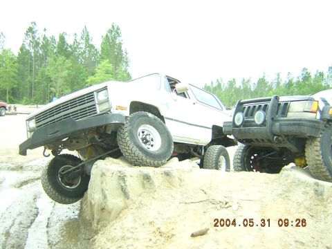 1984 Chevrolet Blazer - Two off-road vehicles navigate a sandy terrain with trees in the background.