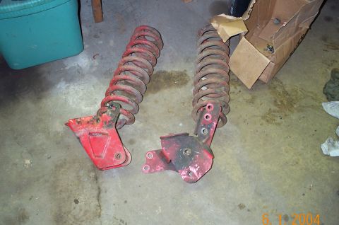 1986 Ford Bronco - A pair of red-painted coil springs still attached to their fabricated frame brackets, laid on a garage floor next to a green tote and a tor…