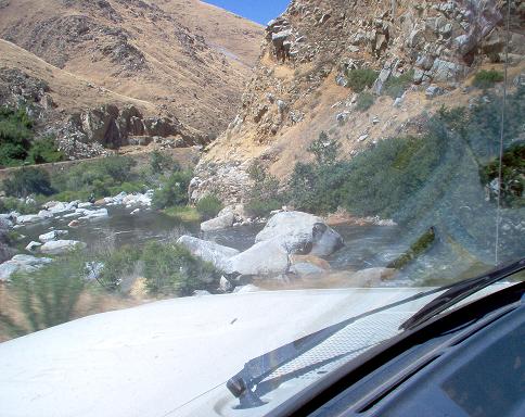 View from inside a vehicle looking out at a rocky riverbed and mountains under a clear blue sky
