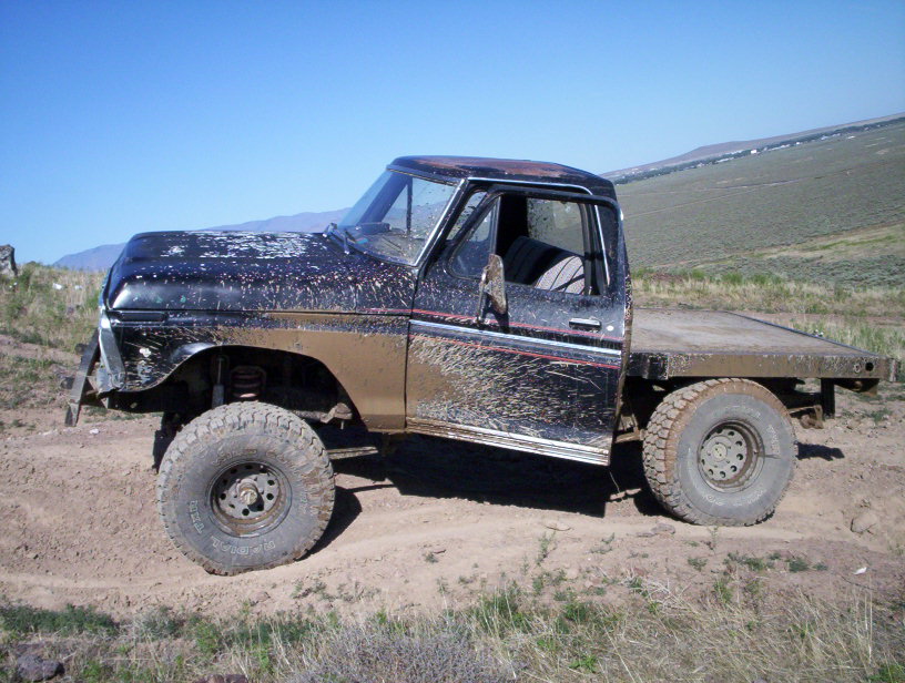 Mud-splattered black Ford F-Series flatbed truck with a lifted suspension and off-road tires, parked on a dirt track
