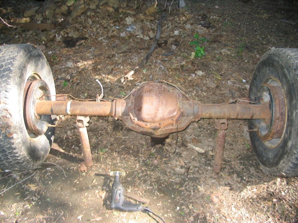 Rusty rear axle assembly with two tires, resting on stands, with a grinder nearby