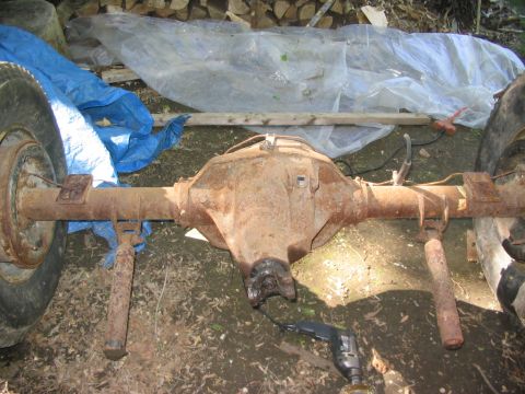 1986 Ford Bronco - A rusty rear axle assembly with tires is shown outdoors, partially covered by tarps.