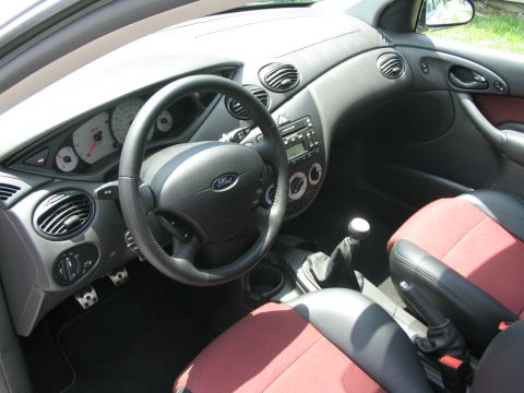2003 Ford Focus - The interior of a 2003 Ford Focus shows a black steering wheel, red and black seats, and the dashboard with a radio and climate controls.