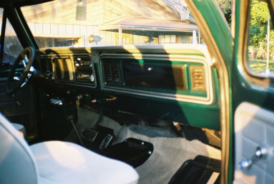 Green Bronco interior through open passenger door showing dash, steering wheel, floor shifter, and white bench seat