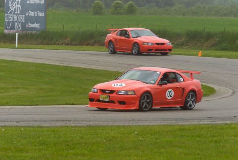 2000 Ford Mustang - Two Ford Mustangs race on a track, with one car prominently in the foreground and another following behind.