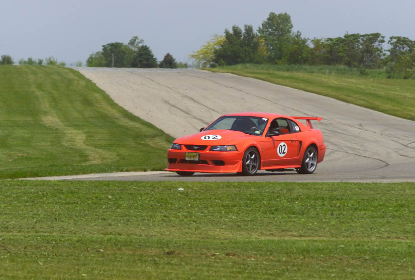 Orange Mustang coupe with "02" roundels and tall rear wing cornering on a road course