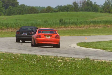 2000 Ford Mustang - An orange 2000 Ford Mustang races on a track behind a black convertible.
