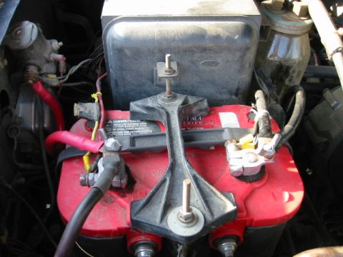 1985 Ford Bronco - Close-up view of a red car battery and its associated wiring and hold-down mechanism within an engine compartment.
