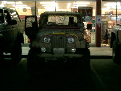 1995 Ford Bronco - A muddy Jeep CJ-5 is parked in front of a store, its driver's side door ajar.
