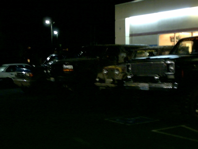 Night parking lot lineup of mud-covered 4x4 trucks and a Jeep outside a lit storefront