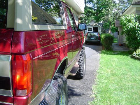 1995 Ford Bronco - This red 1995 Ford Bronco features a tan removable top and is parked on a driveway next to another vehicle.