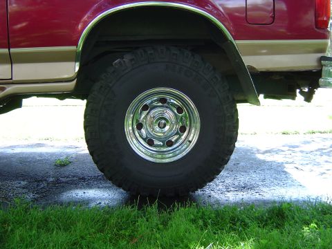 1995 Ford Bronco - Close-up of the rear wheel and fender of a red and tan Ford Bronco, showing the tire tread and chrome rim.