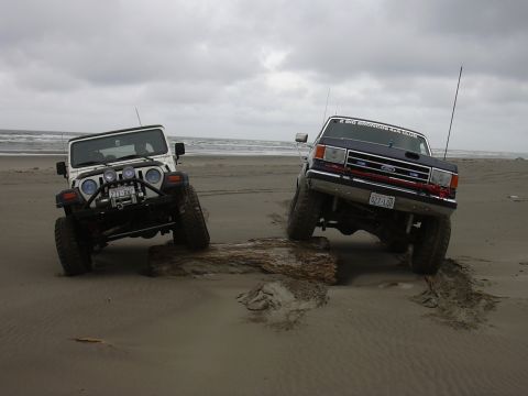 1991 Ford Bronco - Two off-road vehicles navigate a sandy beach, with one vehicle perched on a log.