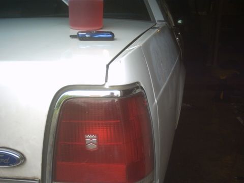 1991 Ford LTD Crown Victoria - Close-up of the right rear corner of a white Ford sedan in a dim garage, showing the chrome-bezeled red tail lamp with a small crest emblem…