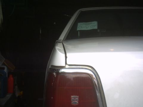 1991 Ford LTD Crown Victoria - Dark garage shot of the left rear corner of a white sedan, showing the chrome-trimmed red tail lamp with a crest badge, the trunk lid edge…