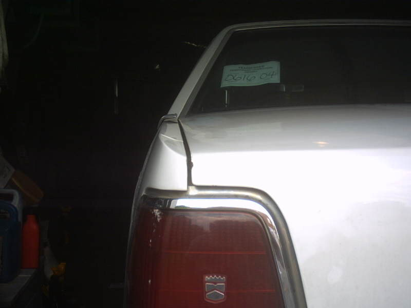 Rear corner of a white sedan with red tail lamp, crest badge and green sticker in the glass