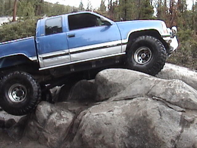 Lifted blue and silver Chevy extended-cab pickup on big off-road tires climbing granite boulders