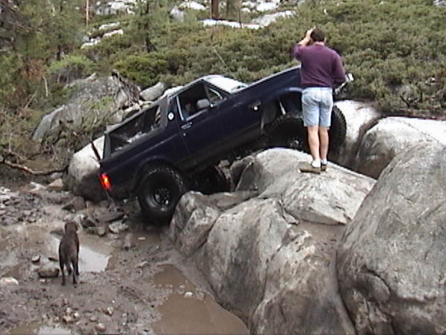Dark blue SUV crawling nose-down off a large boulder on a rocky trail, man and dog nearby