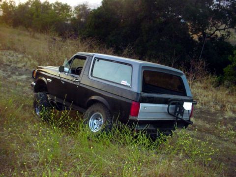 1988 Ford Bronco - This lifted Ford Bronco is parked on a hillside with dry grass and trees in the background.