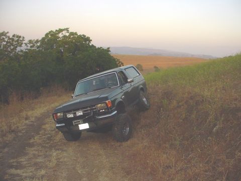 1988 Ford Bronco - A dark SUV navigates an off-road trail with dry grass and rolling hills in the background.