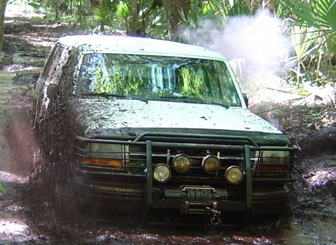 1993 Ford Bronco - Close front view of a mud-splattered 1990s Ford SUV pushing through a muddy trail rut, with mud slung across the hood, windshield and headl…