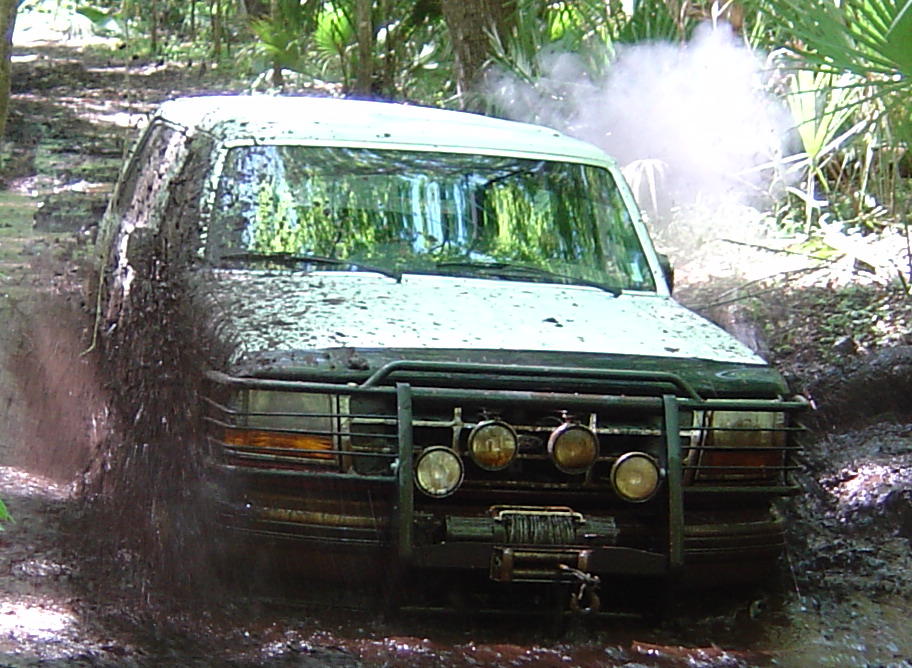 Mud-splattered 1990s Ford SUV with brush guard, four driving lights and winch on a muddy trail