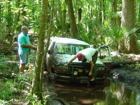 1993 Ford Bronco - A mud-covered white two-door Ford SUV sits nose-down in a flooded rut on a shaded palmetto trail while a man in a red cap leans over the br…