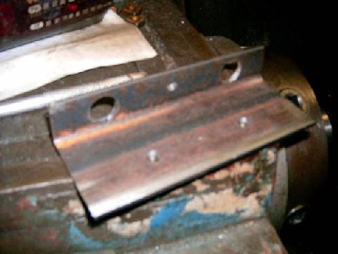 1978 Ford Bronco - A rusty flat steel bracket with two large drilled holes and a row of small punch marks, clamped in a blue bench vise on a cluttered workben…