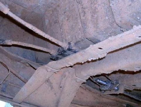 1978 Ford Bronco - Underbody shot looking up at a truck chassis caked in dried tan mud, showing the boxed frame rail, a crossmember, brake or fuel lines and a…