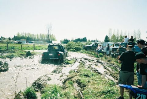 1995 Ford Bronco - A group of people watch as a truck navigates a muddy track on a sunny day.