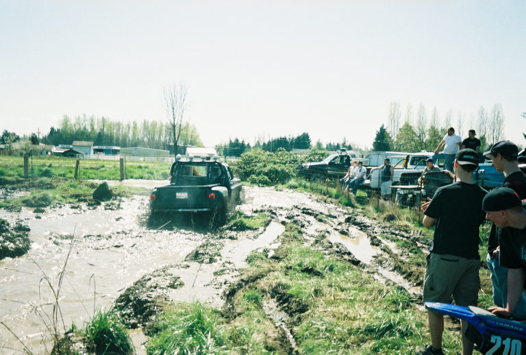 Dark 4x4 crossing a flooded mud-bog pit as spectators watch from the grassy bank