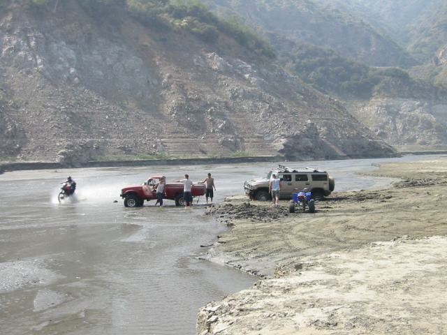Red pickup and tan SUV parked on a wet gravel riverbed with people and an ATV, steep mountain behind