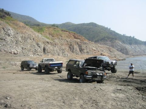 1985 Ford Bronco - Several off-road vehicles are parked on a dirt area next to a body of water with hills in the background.