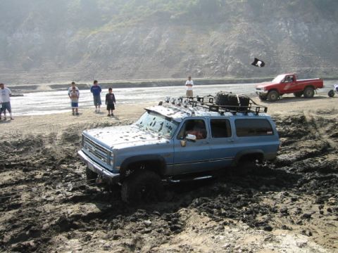 1988 Ford Bronco - A blue SUV is stuck in thick mud while a red pickup truck waits nearby on a dirt track.