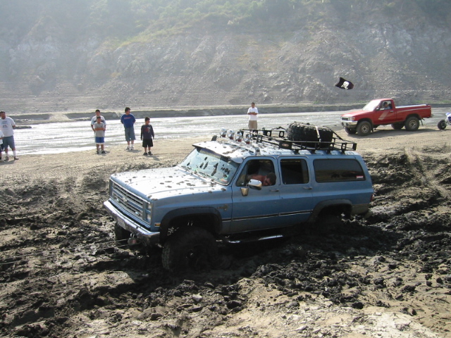 Blue SUV stuck in mud, with a red pickup truck in the background