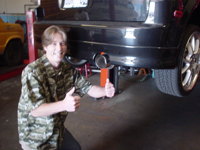 Man in camo shirt giving thumbs up beside a dark SUV's rear bumper and dual exhaust on a shop lift