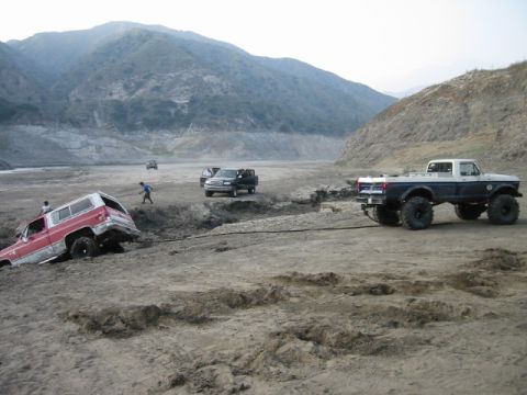 1988 Ford Bronco - A two-tone pickup truck assists a red SUV stuck in a muddy ditch in a desolate, mountainous landscape.