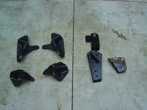 1986 Ford Bronco - Seven black steel mounting brackets laid out on a concrete surface, including several factory-style stamped brackets and one rusty bracket…