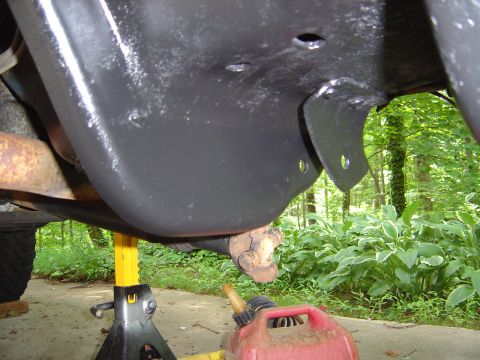 1986 Ford Bronco - Underside view of a vehicle chassis painted flat black, showing a frame crossmember, mounting brackets with bolt holes, and a rusty compone…