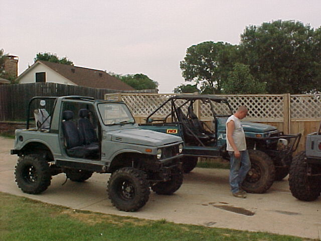 Two modified Suzuki Samurai SUVs parked side-by-side, with a third vehicle partially visible on the right. A man stands between the first two