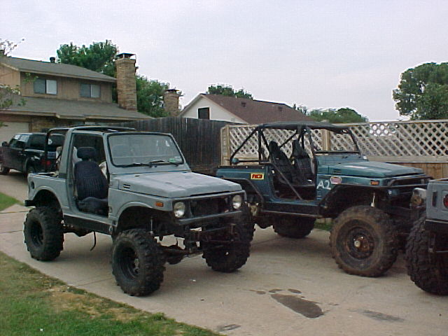 Two modified Suzuki Samurai SUVs parked in a driveway. One is gray with the doors off, the other is teal with a roll cage
