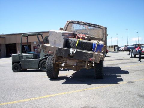1978 Ford Bronco - A heavily used flatbed truck is parked next to a forklift, carrying spare tires and fuel cans.