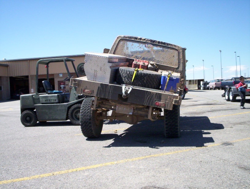 Muddy flatbed truck loaded with tires and gas cans, parked next to a forklift