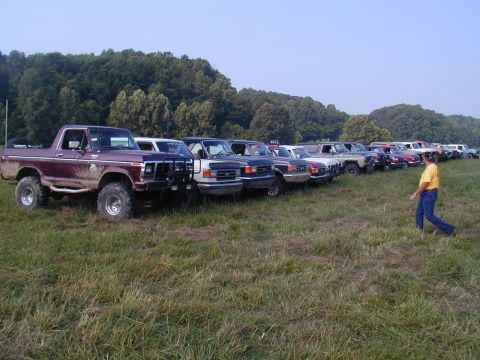 1988 Ford Bronco - A long row of lifted Ford Broncos and full-size trucks parked nose-out in a grassy field at an off-road club gathering, with wooded hills b…