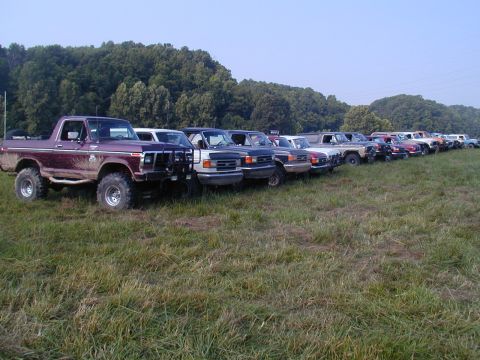 1988 Ford Bronco - A line of SUVs and trucks are parked in a grassy field under a clear sky.