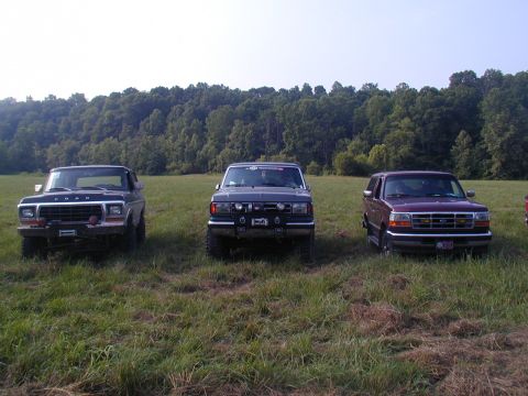 1988 Ford Bronco - Three Ford SUVs are parked in a grassy field with trees in the background.