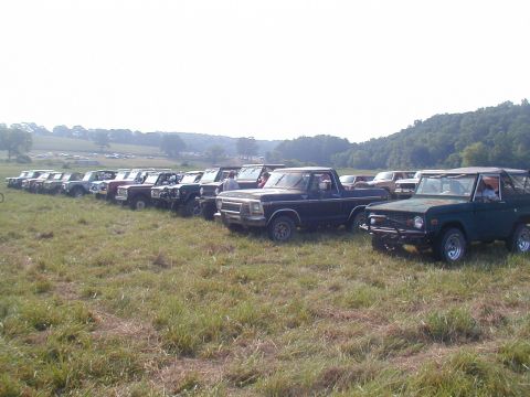 1988 Ford Bronco - A line of trucks and SUVs are parked in a grassy field under a hazy sky.