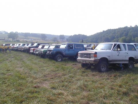 1988 Ford Bronco - A line of off-road vehicles, including a white 1987 Ford Bronco, are parked in a grassy field on a sunny day.