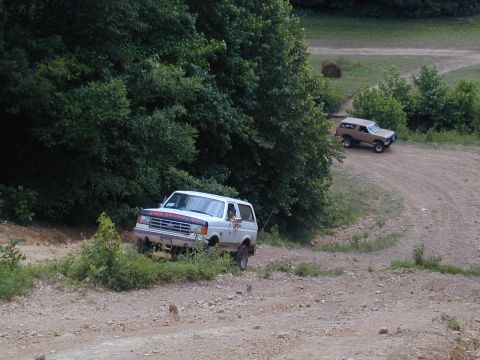 1988 Ford Bronco - Two Ford Broncos navigate a dirt trail surrounded by lush green trees and foliage.