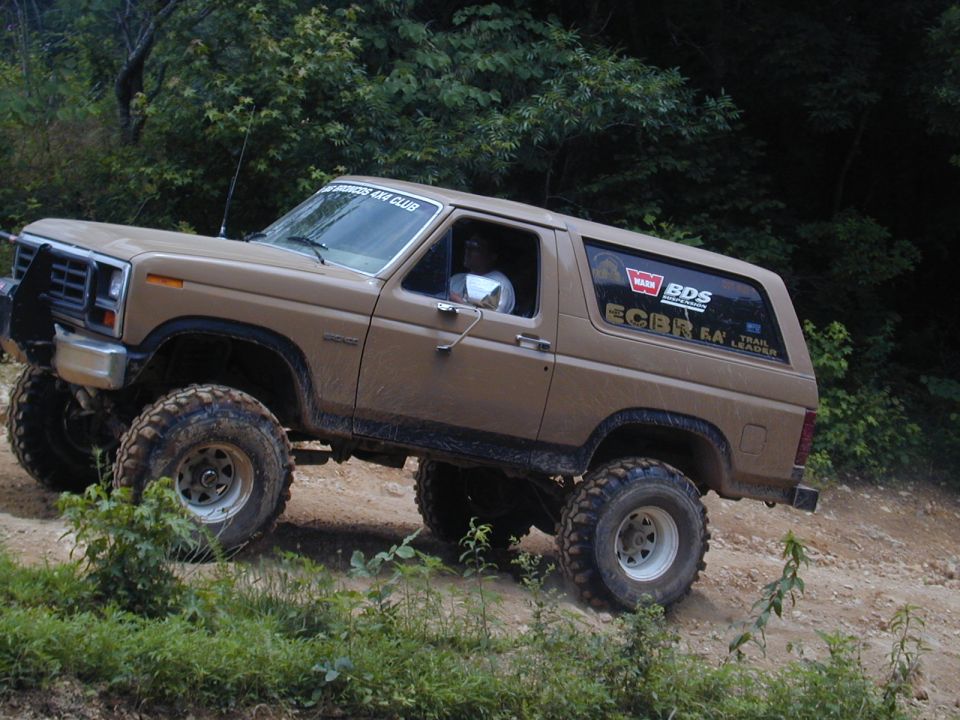 Tan 1988 Ford Bronco with lifted suspension and off-road tires driving on a dirt trail